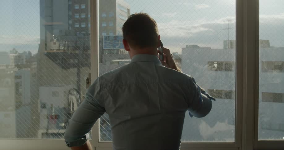 Man stand at window overlooking downtown talking on phone