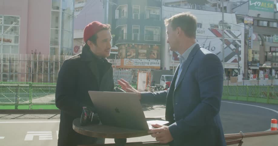A man shows a person something on a laptop near a city street