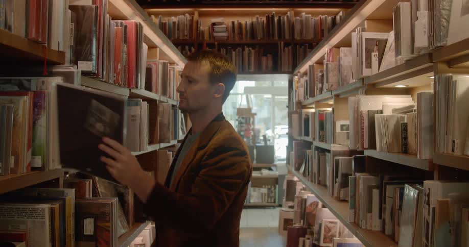 A man walking through an aisle in a library before talking a book from a shelf and looking through it