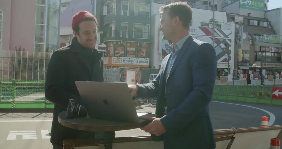 Two men stand outside and talk and look at a laptop