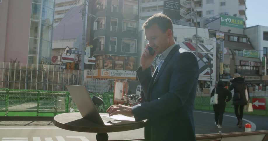 A man works on his laptop outside as people walk down a city pathway
