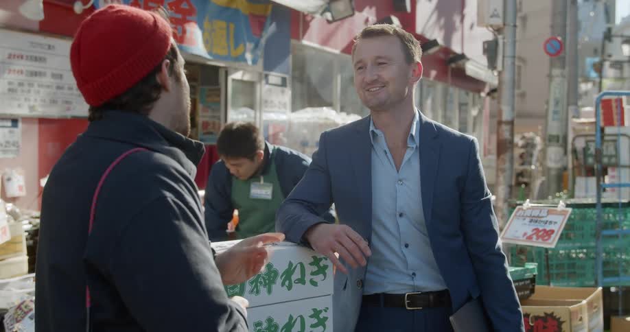 Two men stand at a market and chat with the worker behind them