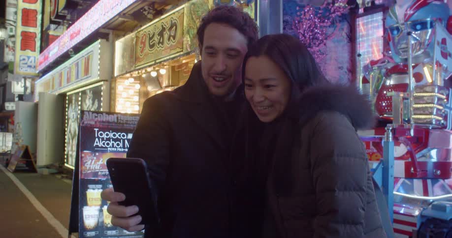 A man and woman pose together for a selfie in front of a store with flashing lights on a city street at night