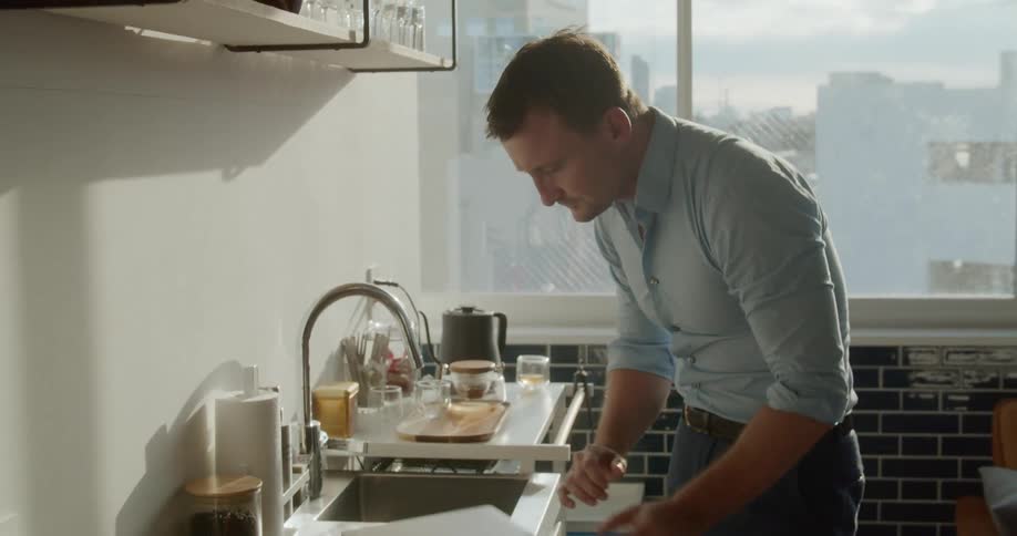 A man typing on a laptop next to a sink before closing it and carrying it away