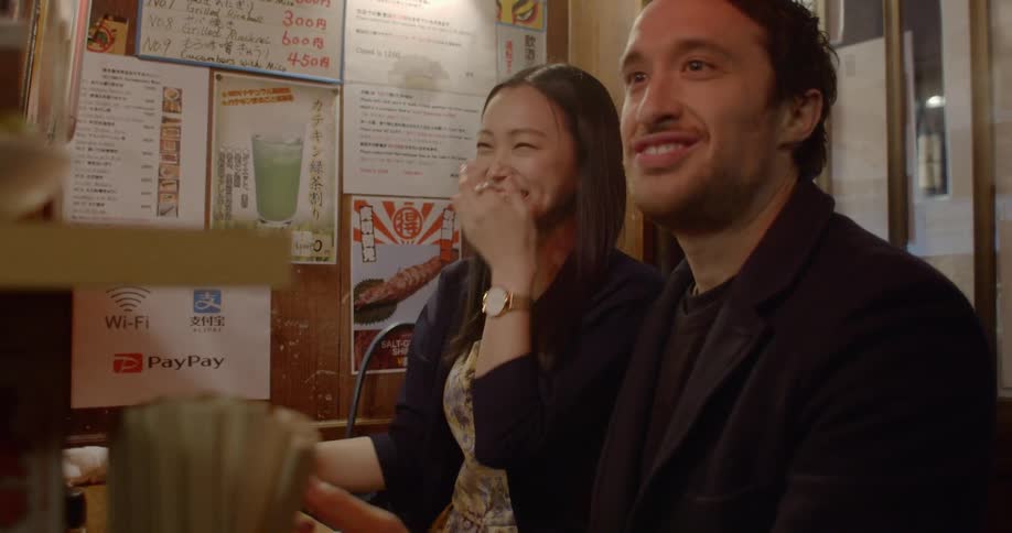 A woman laughs as she accepts a drink over the counter while she and her date sit at a sushi bar