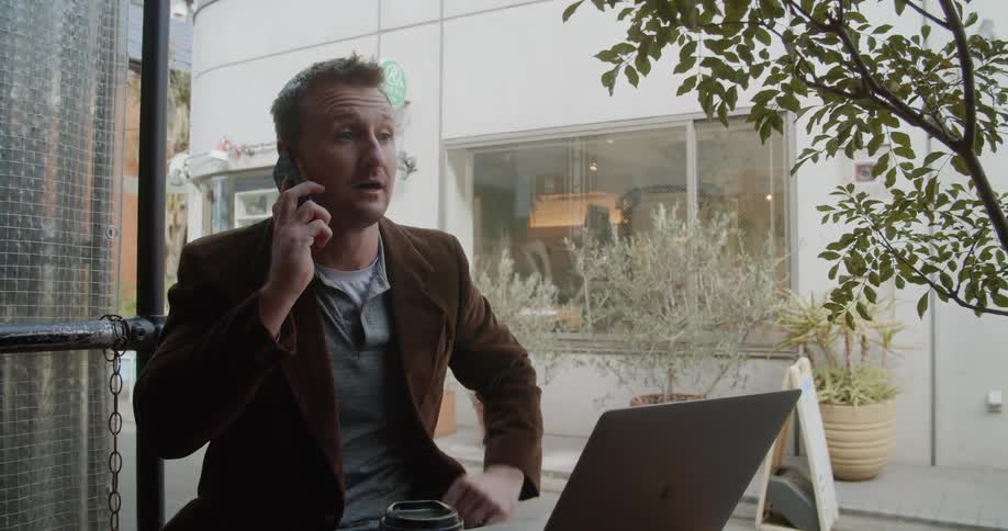 A man sits at an outdoor cafe talking on the phone and with his laptop open