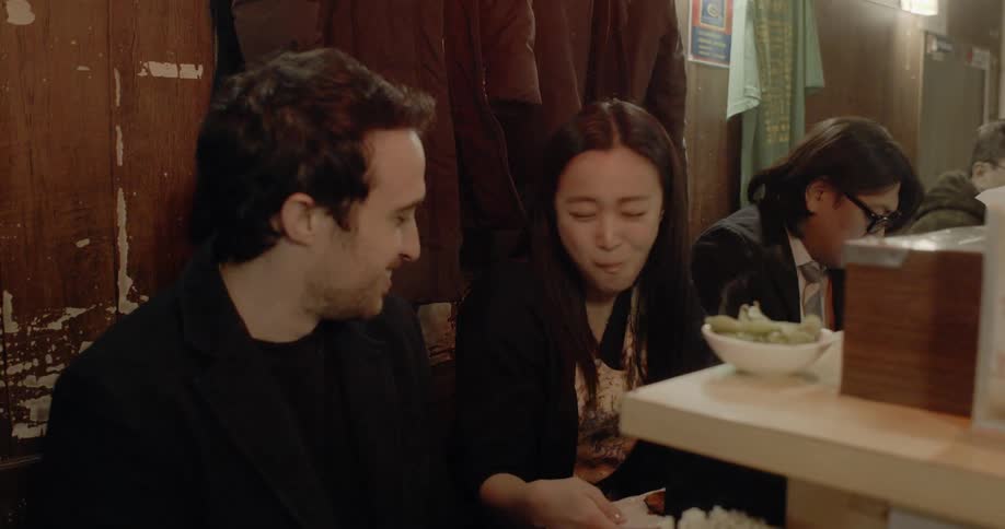 An asian woman eats in a restaurant while a white man watches her