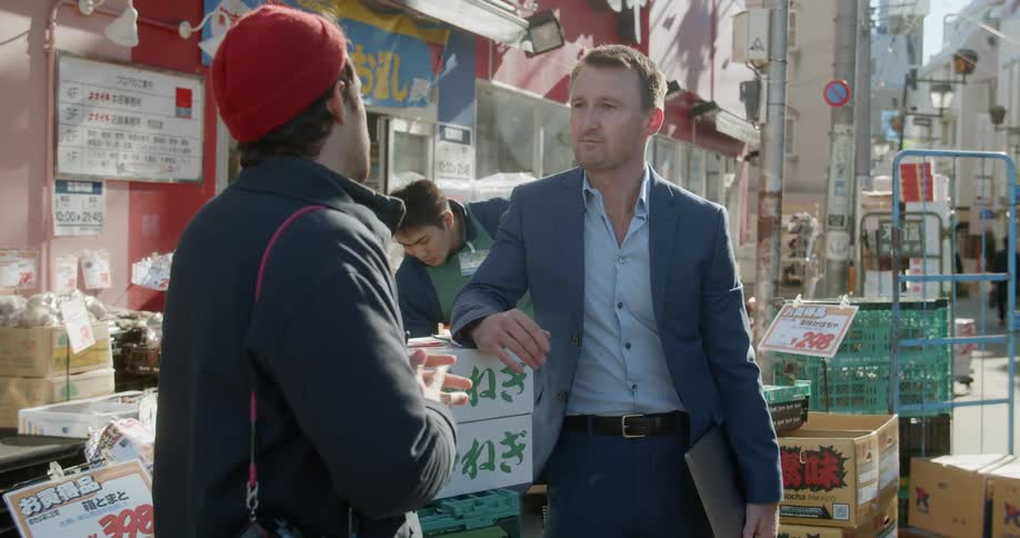 A man leans on a box as he smiles and talks to another at an outdoor asian market