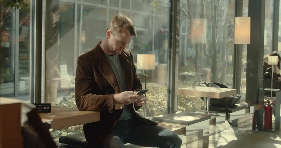 A man sits on a bench beside a window on his cell phone