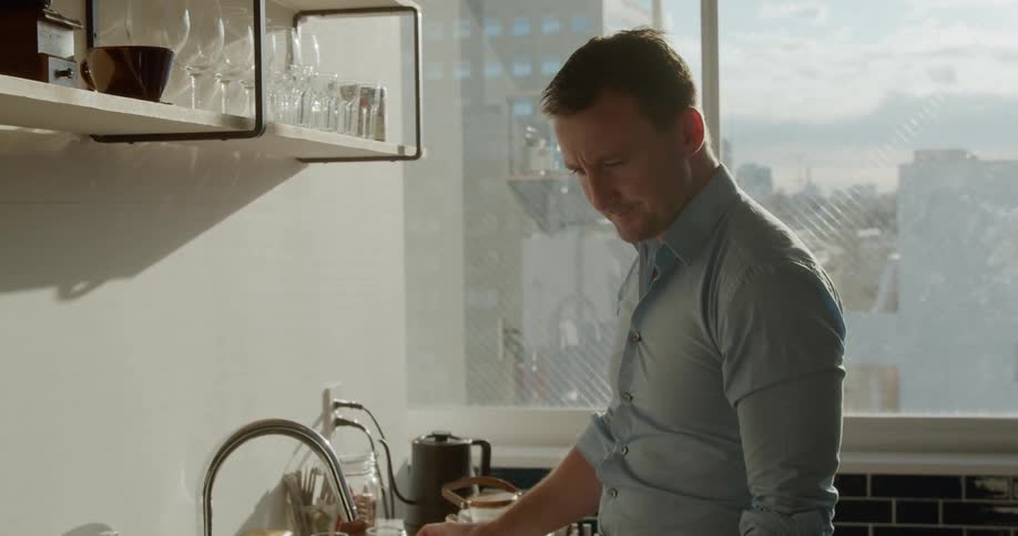 A man having a drink while standing near a faucet and typing on a laptop on the counter