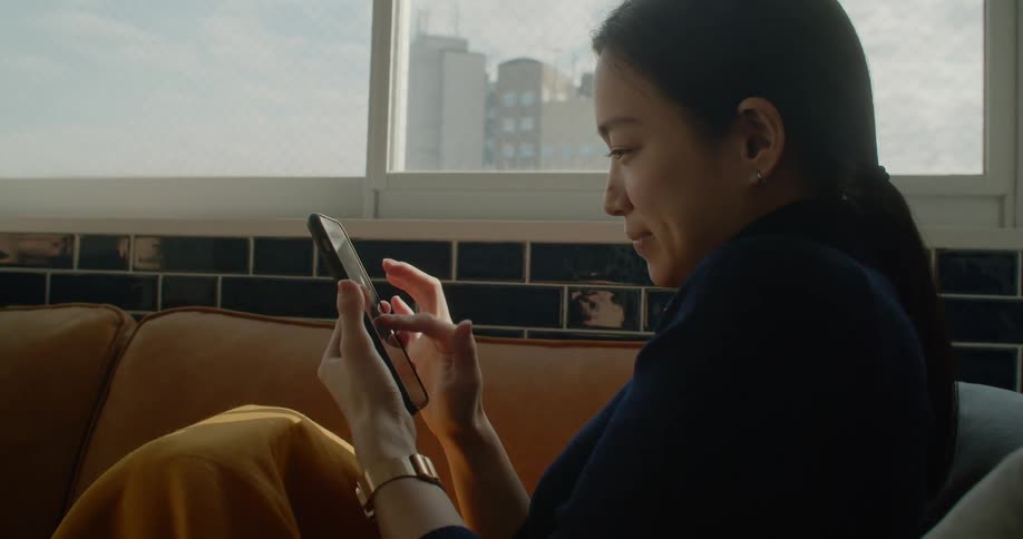 A woman sits on a couch with a cell phone and then shows someone else the screen