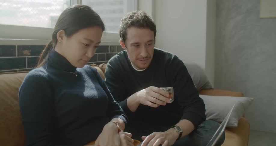 White man and asian woman sitting on the couch uses a laptop in the living room