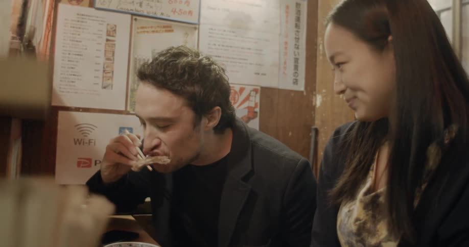 An asian woman watching as a white man tries food in an asian restaurant