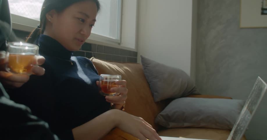 Asian woman and white man sitting on the sofa uses a laptop and drink tea at home