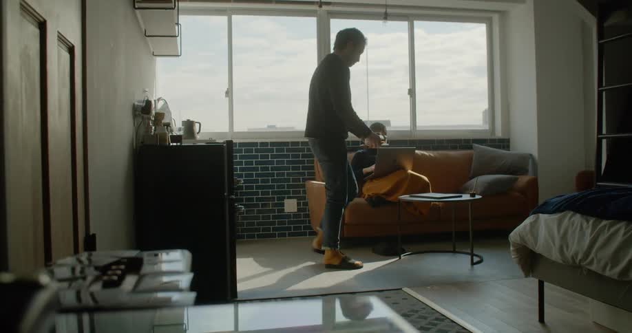 A man puts a tray with drinks on a coffee table while a woman uses a laptop on a sofa
