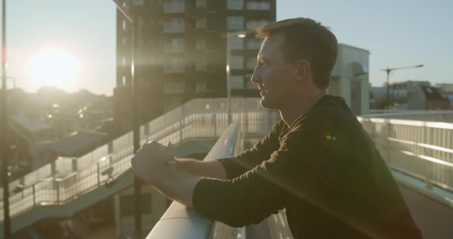 Man resting his elbows on the steel railing stands on the overpass at sunset