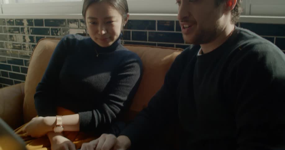 A man smiles as he types on his laptop while sitting on a couch next to a woman