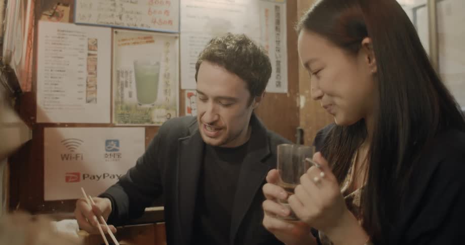 A woman holding a beer glass watches as a man picks up a piece of food using chopsticks and takes a bite