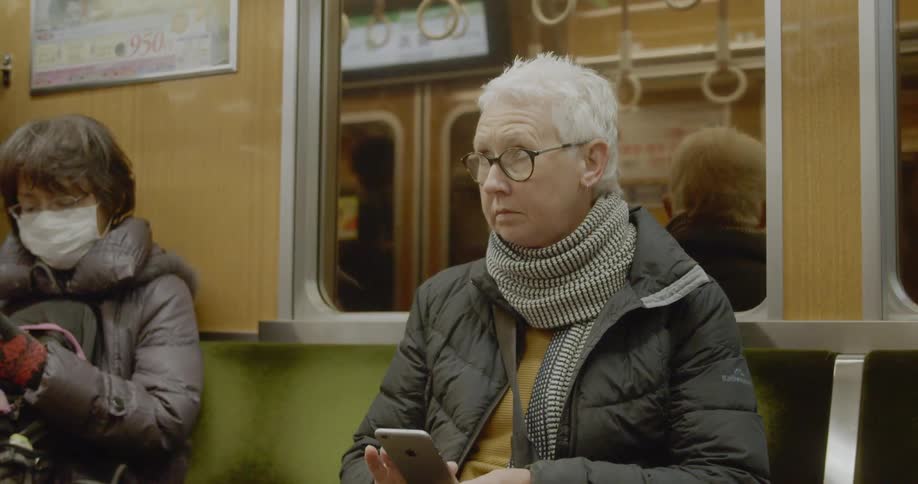 A woman uses her smartphone while riding a subway train and sitting next to a masked passenger