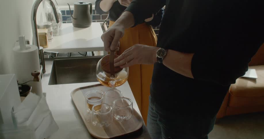A man pouring tea in the kitchen while a woman in the background is on a smart phone