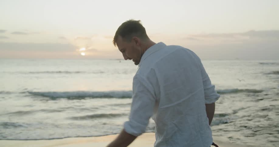Man and his dog stands on the sand of a beach looking at the sunset