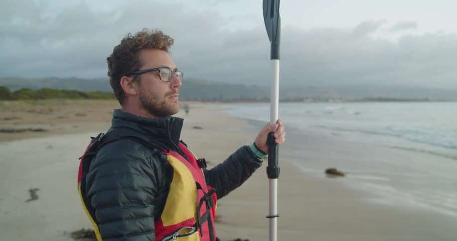 Man wearing life jacket stands on a beach holding an oar