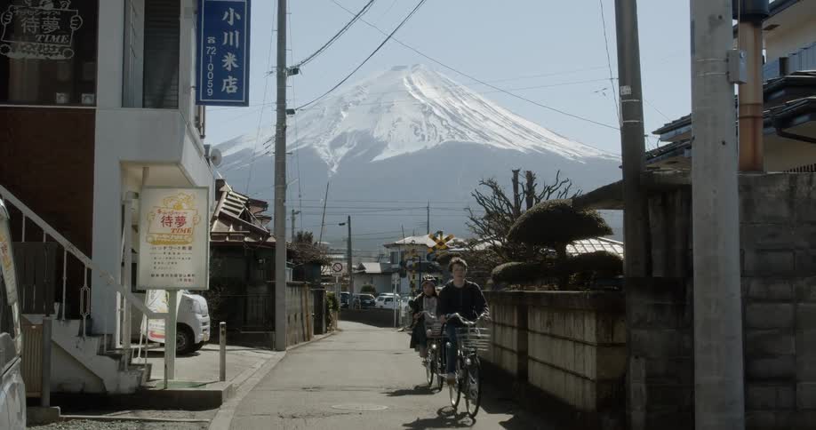 People ride bicycles along street between buildings with mount fuji in the background
