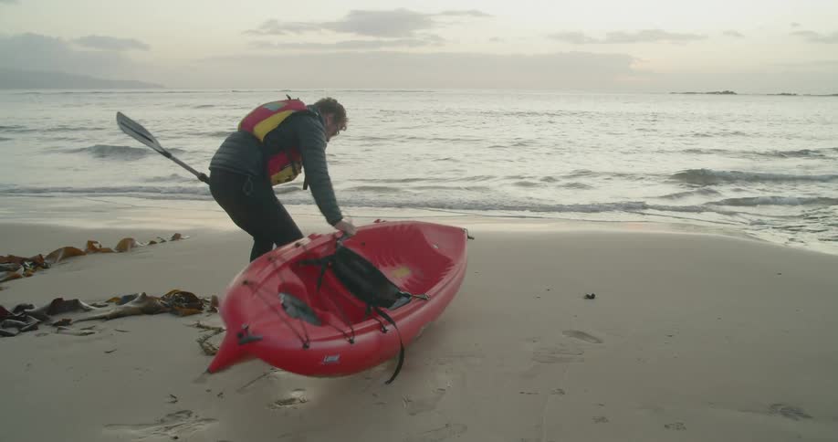 A man carries his kayak towards the water and then sets it down in the sand