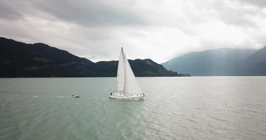 Small boat follows in wake of sailboat with fog covered mountains on the horizon