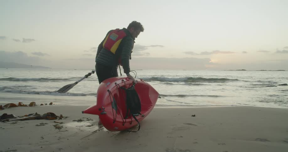 A white man carries a kayak and oar across the beach