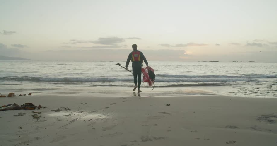 Man walks toward surf carrying paddle and small boat