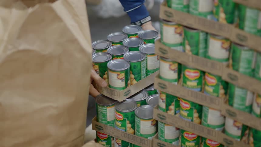 A white man lifts a box of canned goods off of a stack of product