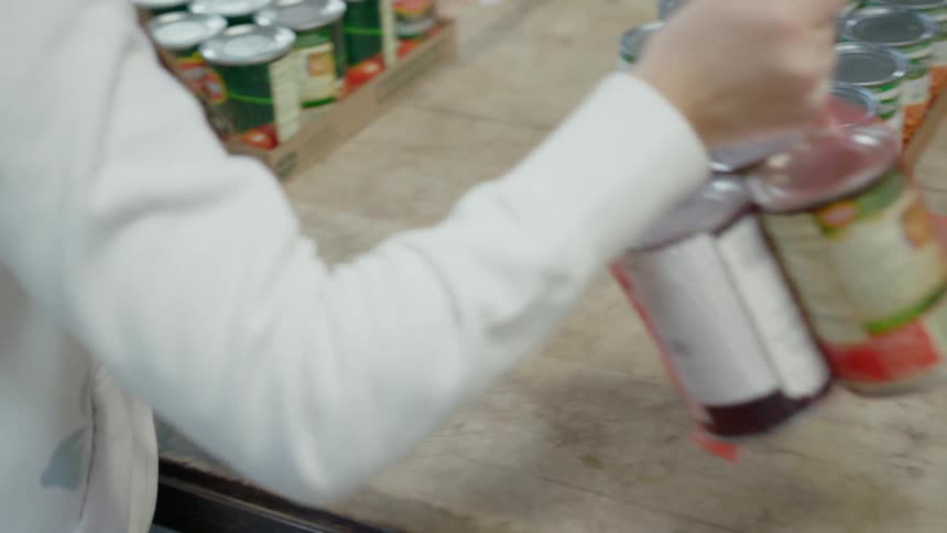 Female volunteering at a food pantry picks up four canned goods that are contained in netting material and moves them to the other end of the table