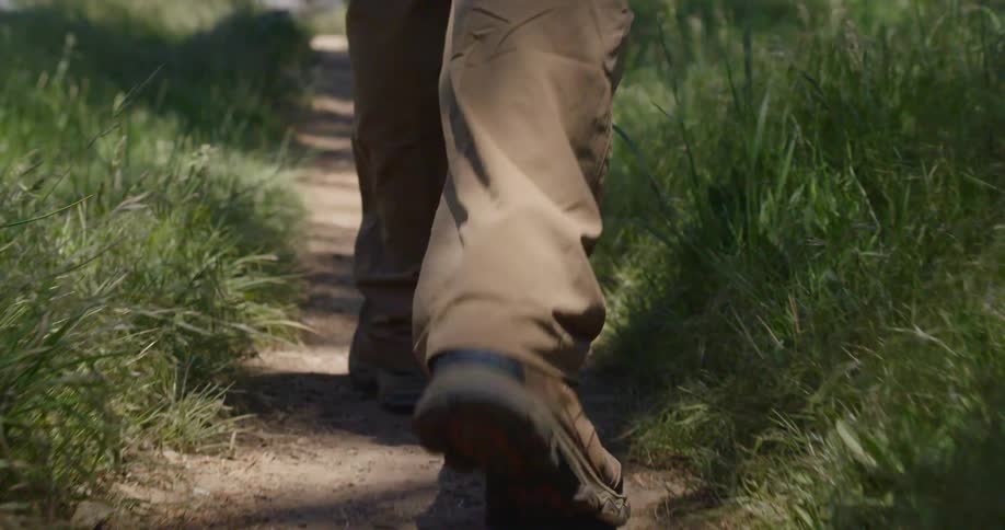 The orange and black soles and treads of a persons hiking boots as the walk on a narrow dirt trail between the grass