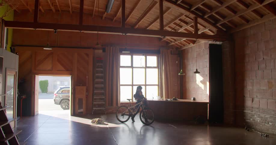 A woman wheels her bike into a building and drops her backpack on the floor