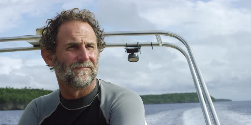A middle-aged man with a graying beard sitting on a boat on a partially cloudy day