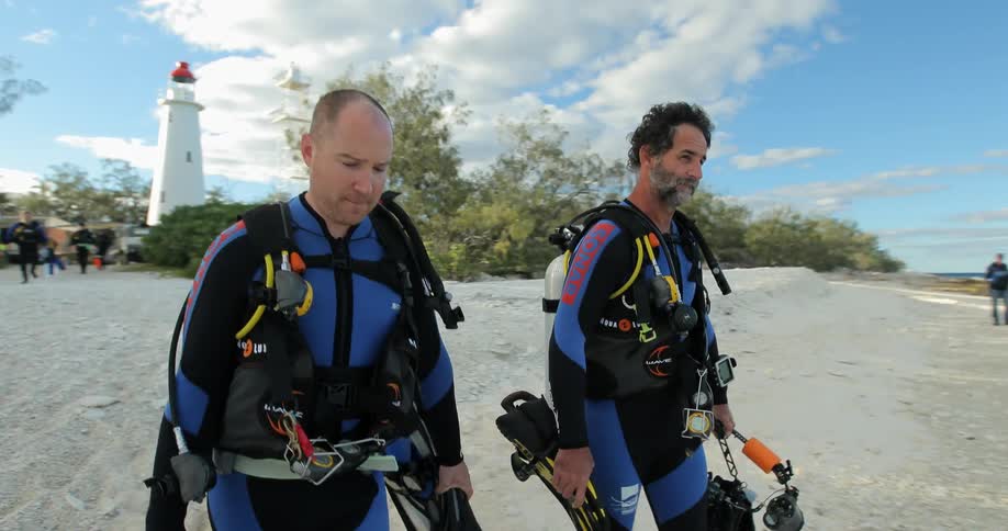 Two men dressed in wet suits and carrying diving gear walking down a beach towards the ocean