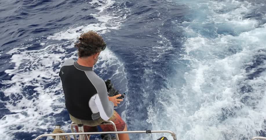 A man sitting on the edge of a boat grabs a camera and puts on his goggles