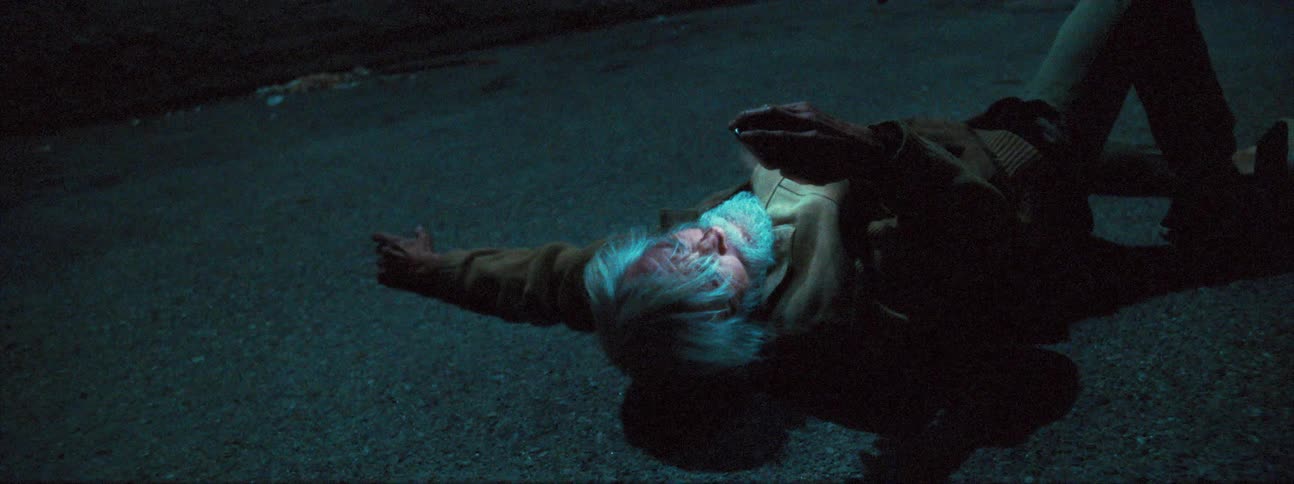 An elderly man lies on the ground outside at night and uses his cell phone