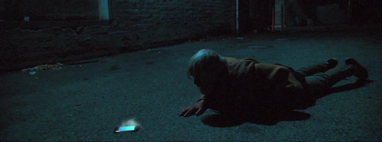 A man falls to the ground and crawls toward a cell phone that is flashing and smoking