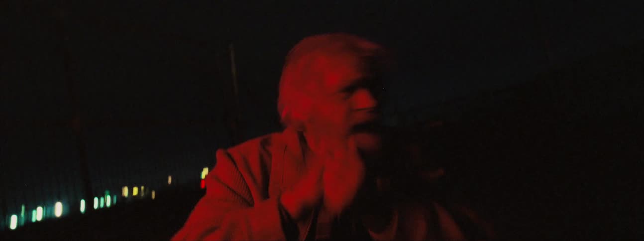 Elderly man sits outdoors on ground at night rubbing face and beard