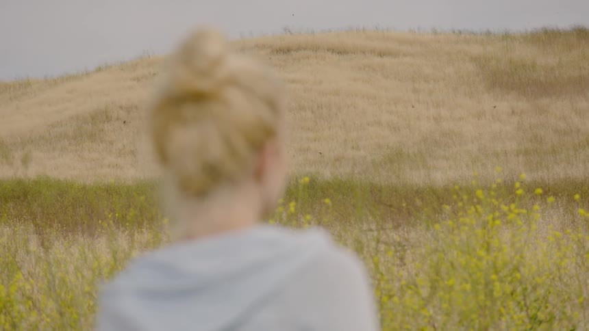 A woman looks around an open field as birds fly by