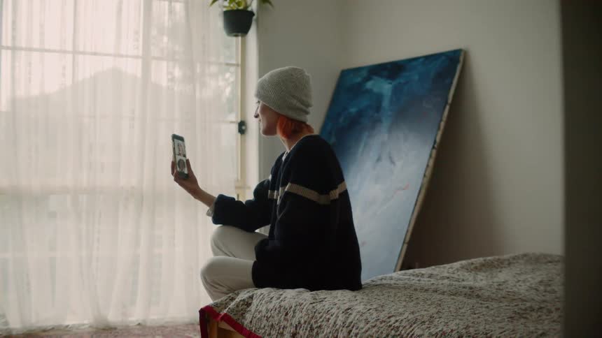 Woman wearing a toque sits on the edge of a bed as she video chats with her cell phone