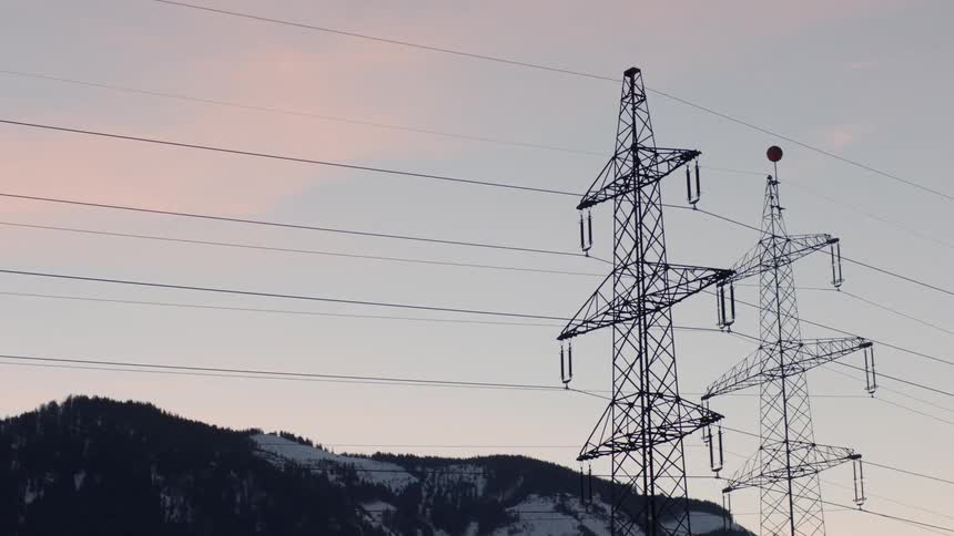 Power lines from transmission towers against the sky with snow dotted mountain slopes behind