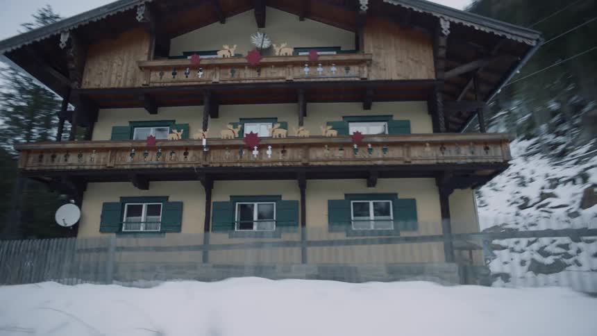 Road passes by three story inn standing in valley between forest and snow covered mountains