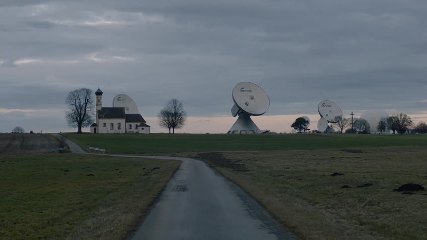 Road leads past mowed fields to building and radio telescopes under a cloudy sky