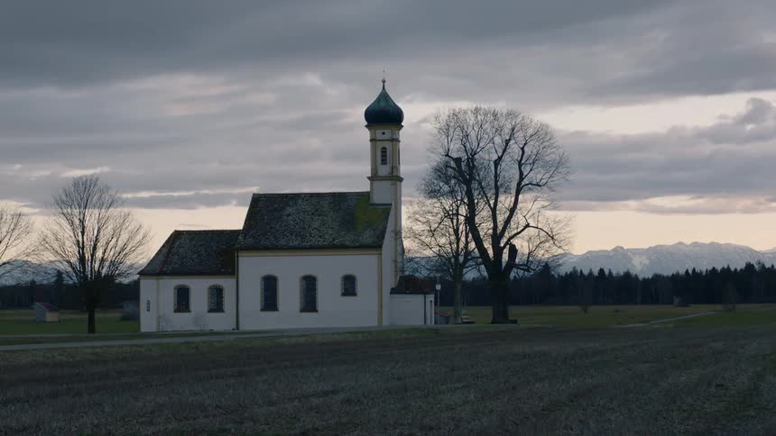 A church with a dusting of snow on the roof sits alone in a field with bare branch trees around it