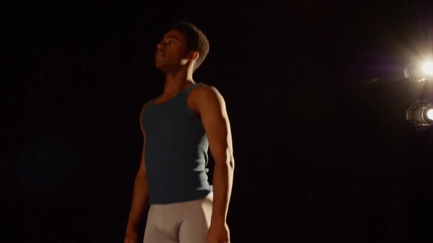 A young man wearing tights and a tank top does ballet leaps while a bright light shines