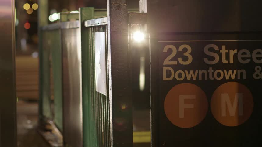 Car headlights shine on the sign and railing of a downtown subway station