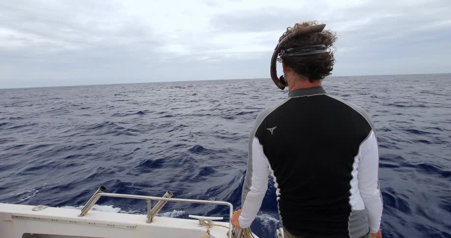 A man wearing snorkeling gear is standing on the edge of a moving boat